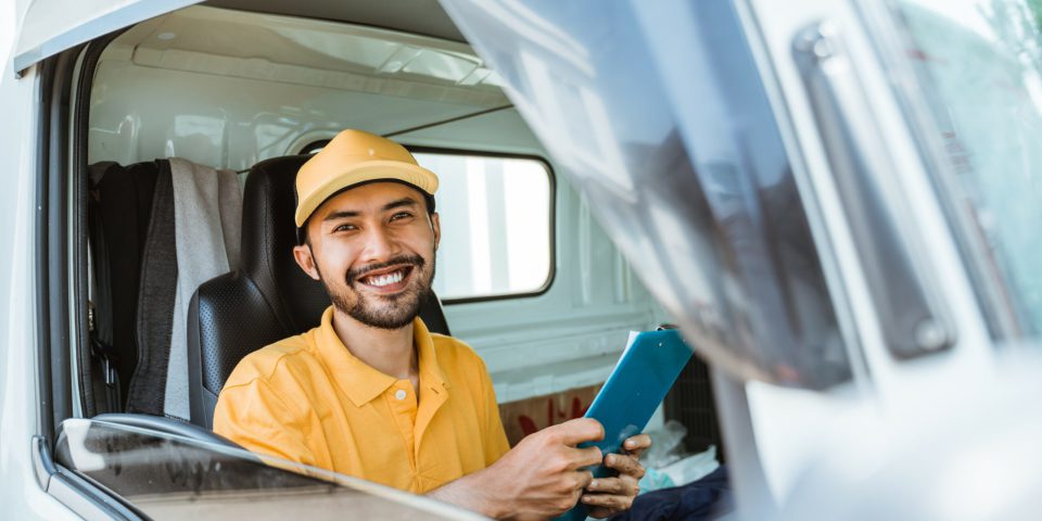 delivery man in yellow uniform smiling holding clipboard sitting while driving goods truck
